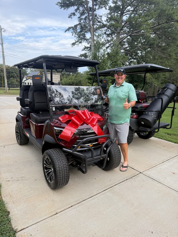 A man beside a new red lifted golf cart wrapped with a large red bow in a driveway