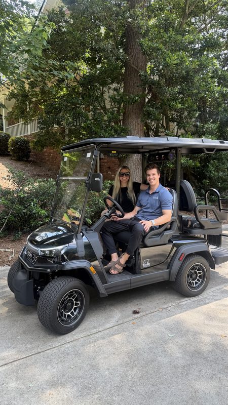 A couple seated in their new black lifted golf cart on a shaded driveway